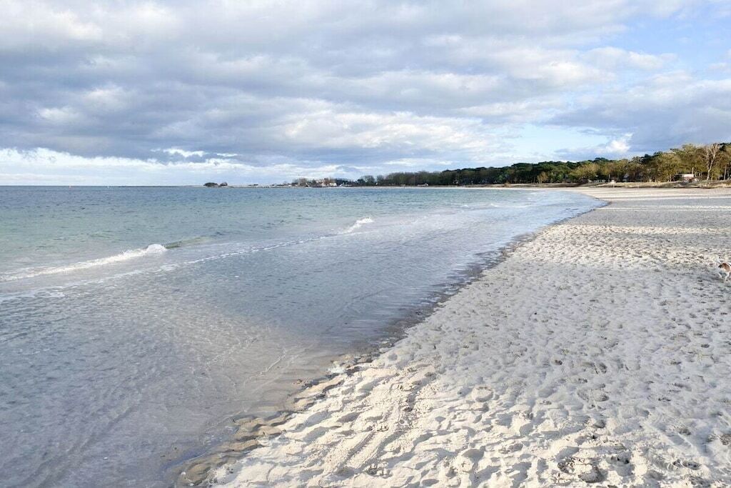 A beach with clear water, a sandy shore, and a distant shoreline with trees and buildings.