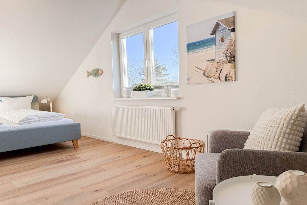 A bedroom with a bed, a window, a radiator, a wicker basket, and a painting of a beach scene.