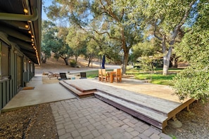 A deck with outdoor seating, a pathway leading to a building, and trees in the background. - Rancho de los Venados (Santa Ynez)