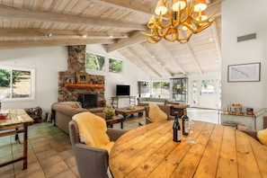 A spacious living area with a stone fireplace, wooden ceiling beams, and a round wooden table with wine glasses and a bottle. - Rancho de los Venados (Santa Ynez)