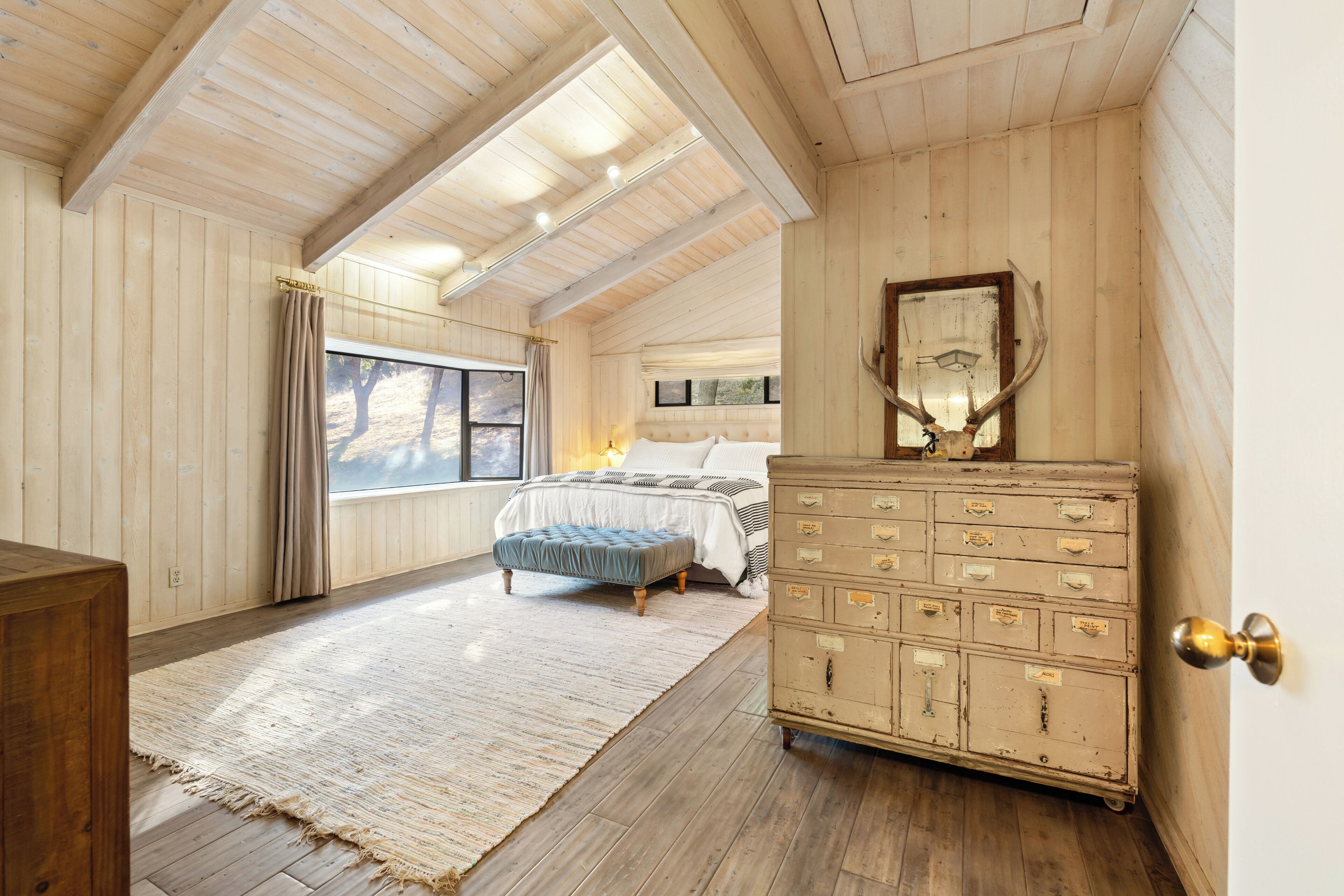 A bedroom with a large window, a bed, a dresser, and a mirror.