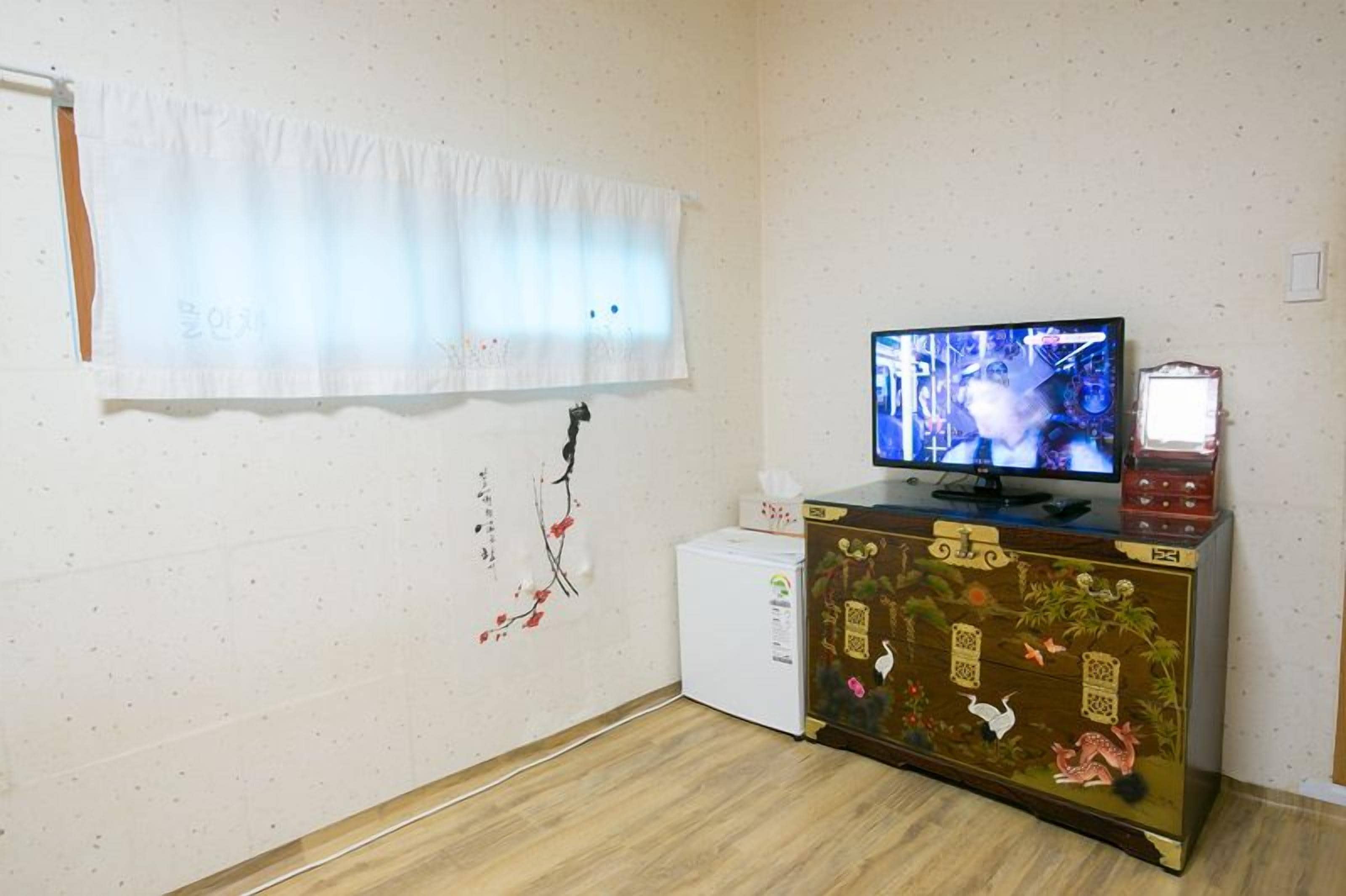 Una habitación con televisor en un mueble, un refrigerador pequeño y una ventana con cortina.