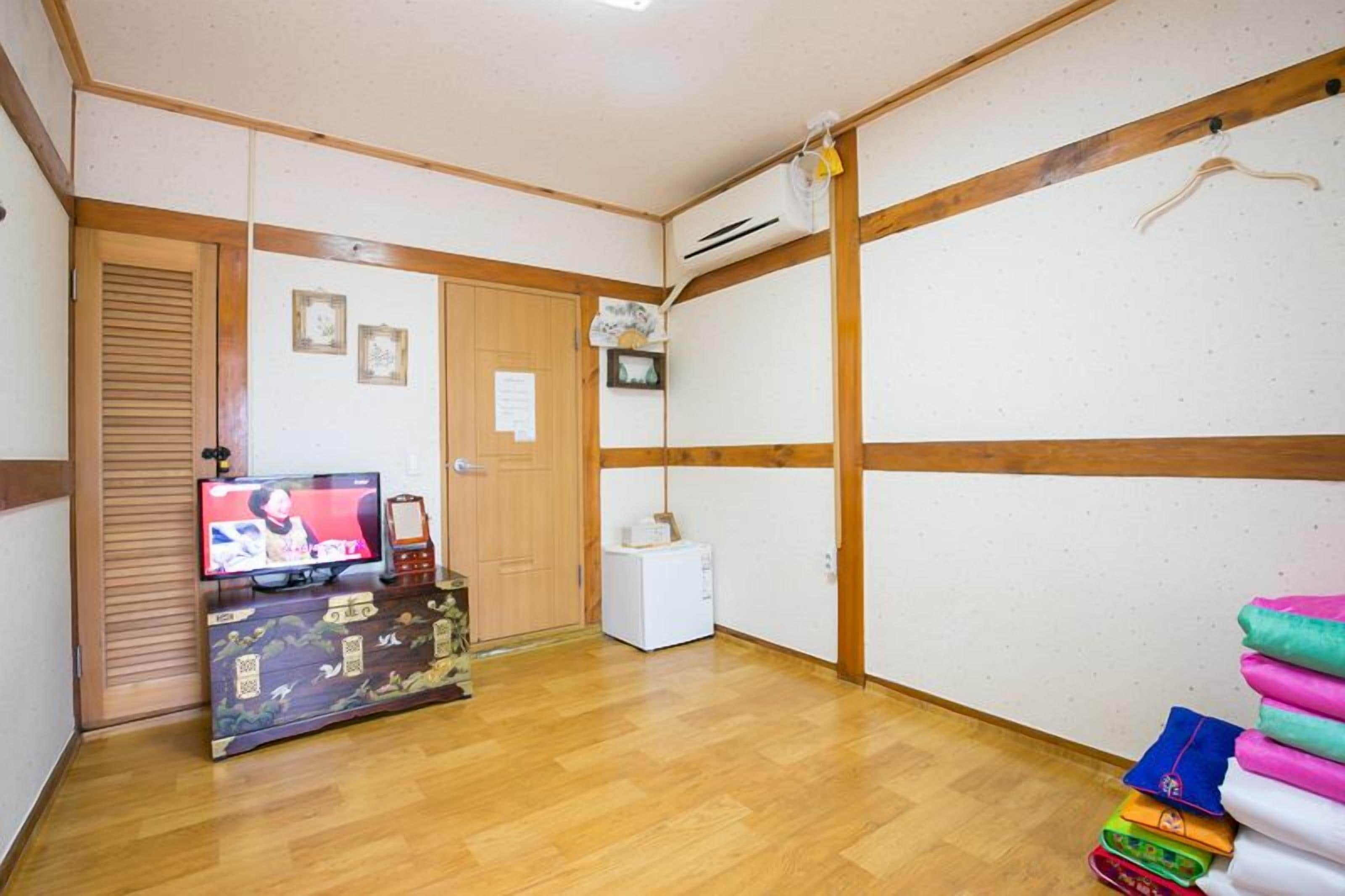 Una habitación con piso de madera, un televisor sobre un mueble bajo, un electrodoméstico blanco pequeño y una pila de toallas dobladas.