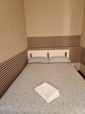 Un letto singolo con una coperta a motivi e cuscini, un asciugamano bianco piegato e un poggiatesta a righe.
