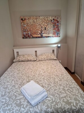 Un llit ben arreglat amb un estovat estampat, un penjall decoratiu a la paret i una tovallola blanca doblegada sobre el llit.