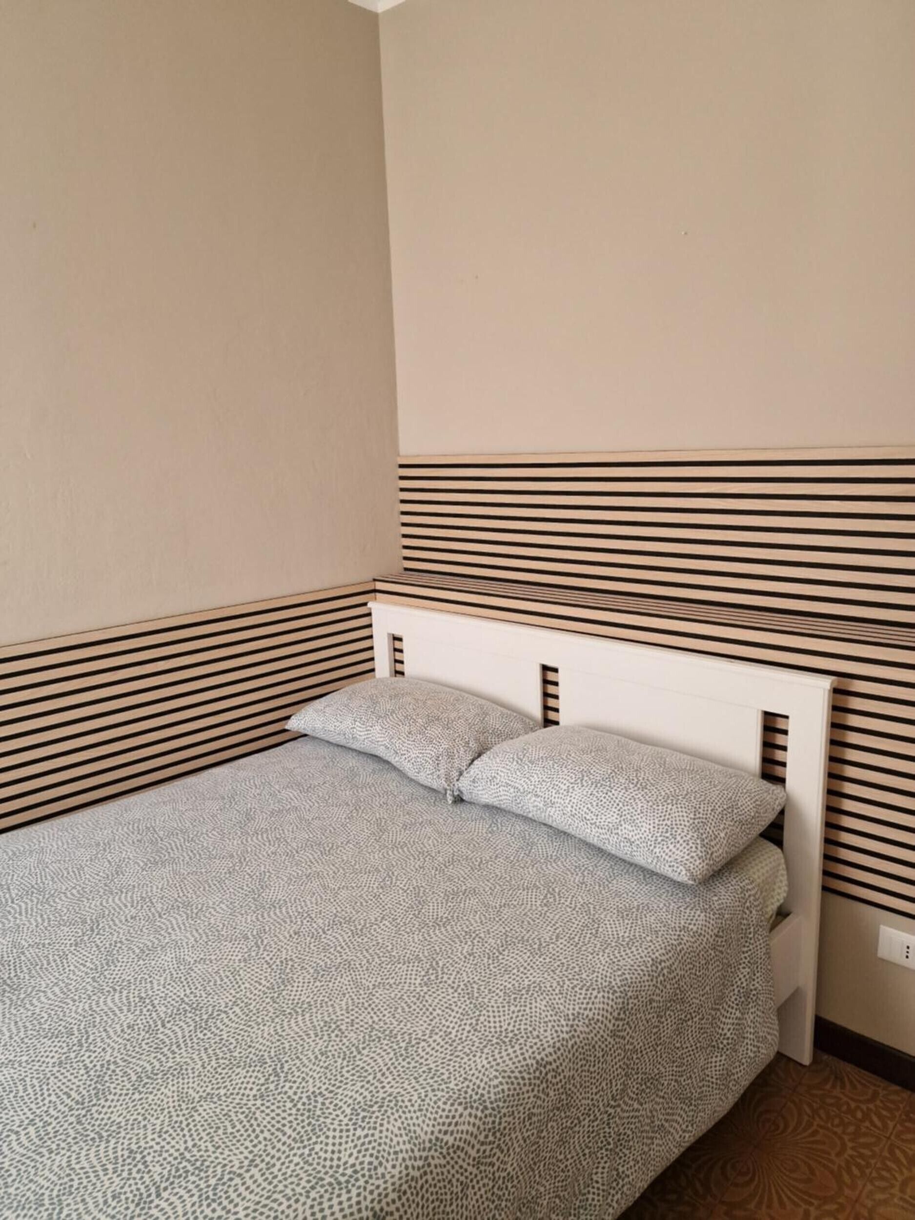 Un letto singolo con una coperta a motivi e due cuscini, appoggiato contro un muro a righe.