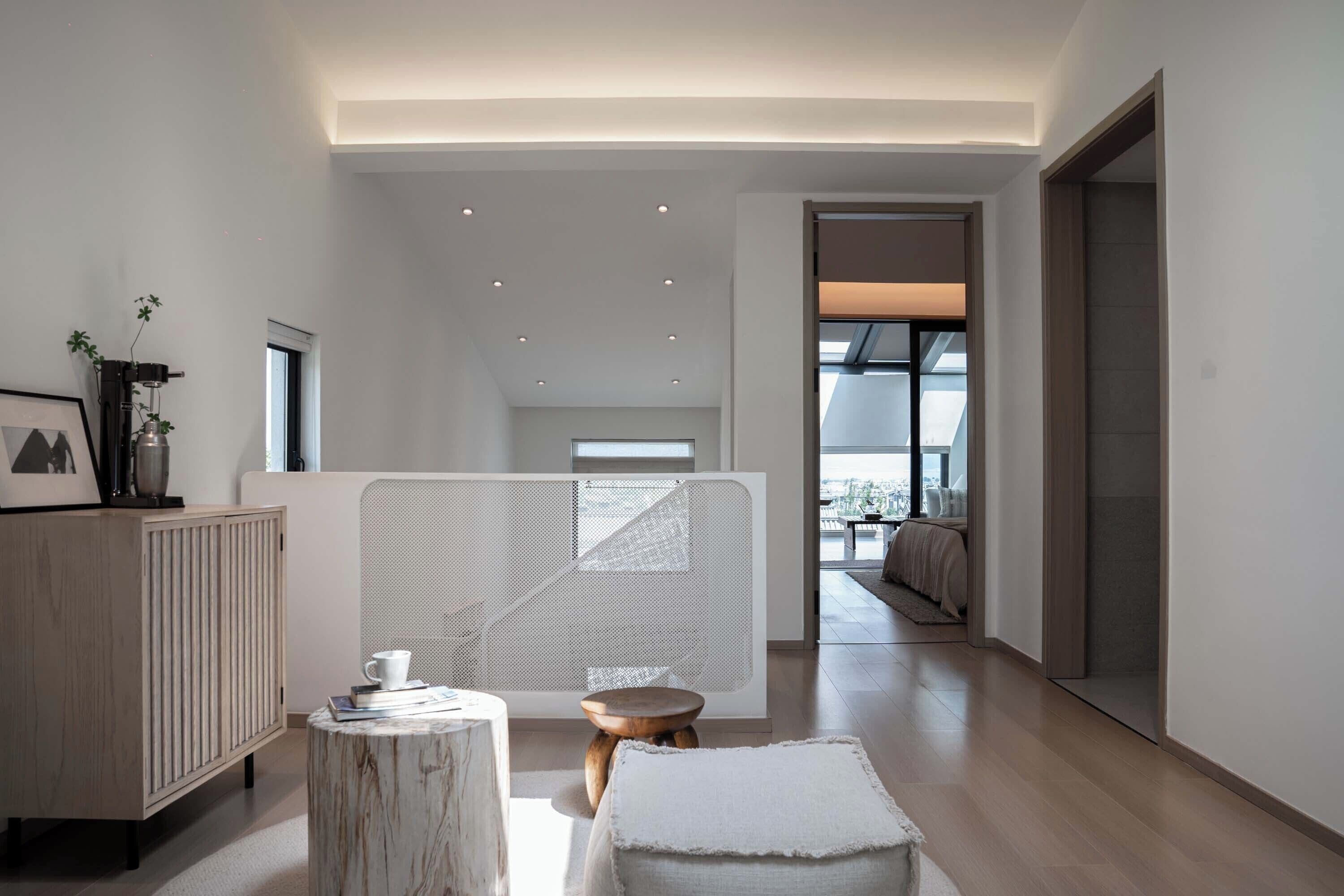 Un interior moderno con un mueble de madera, un taburete redondo de madera y un pouf blanco.