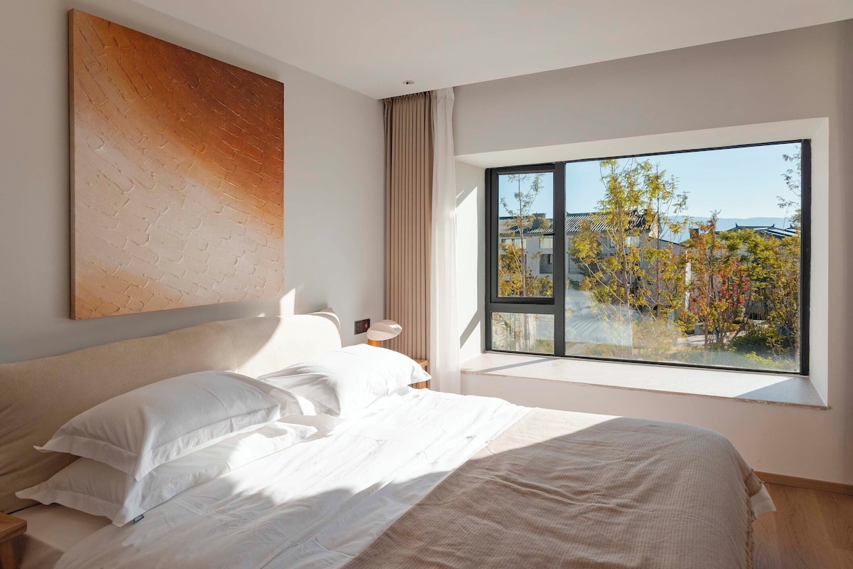 Quarto moderno com uma janela grande, cama com roupa de cama branca e uma obra de arte de parede texturizada.