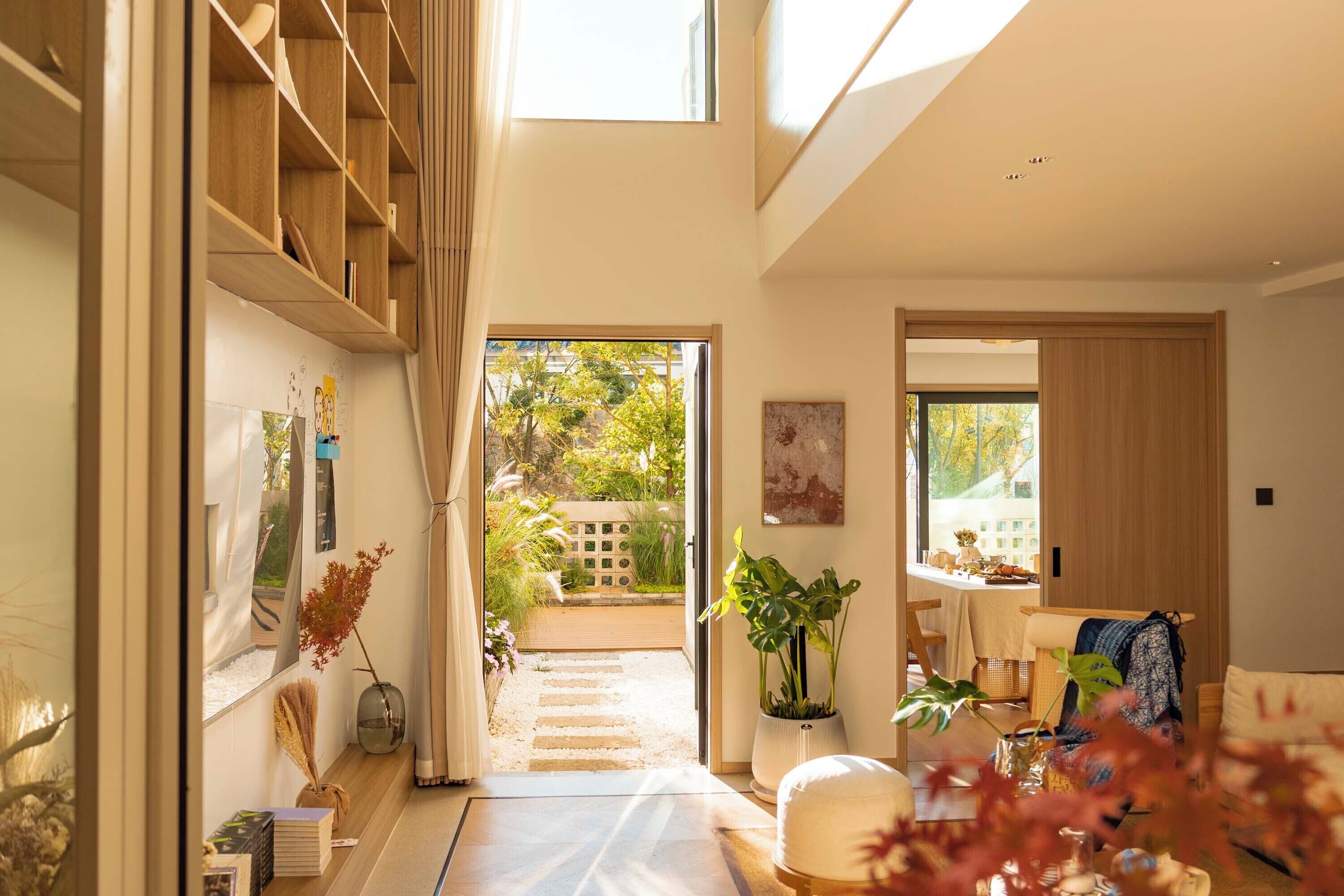 A spacious room with a wooden bookshelf, a dining area with a table and chairs, and a view of a garden through a glass door.