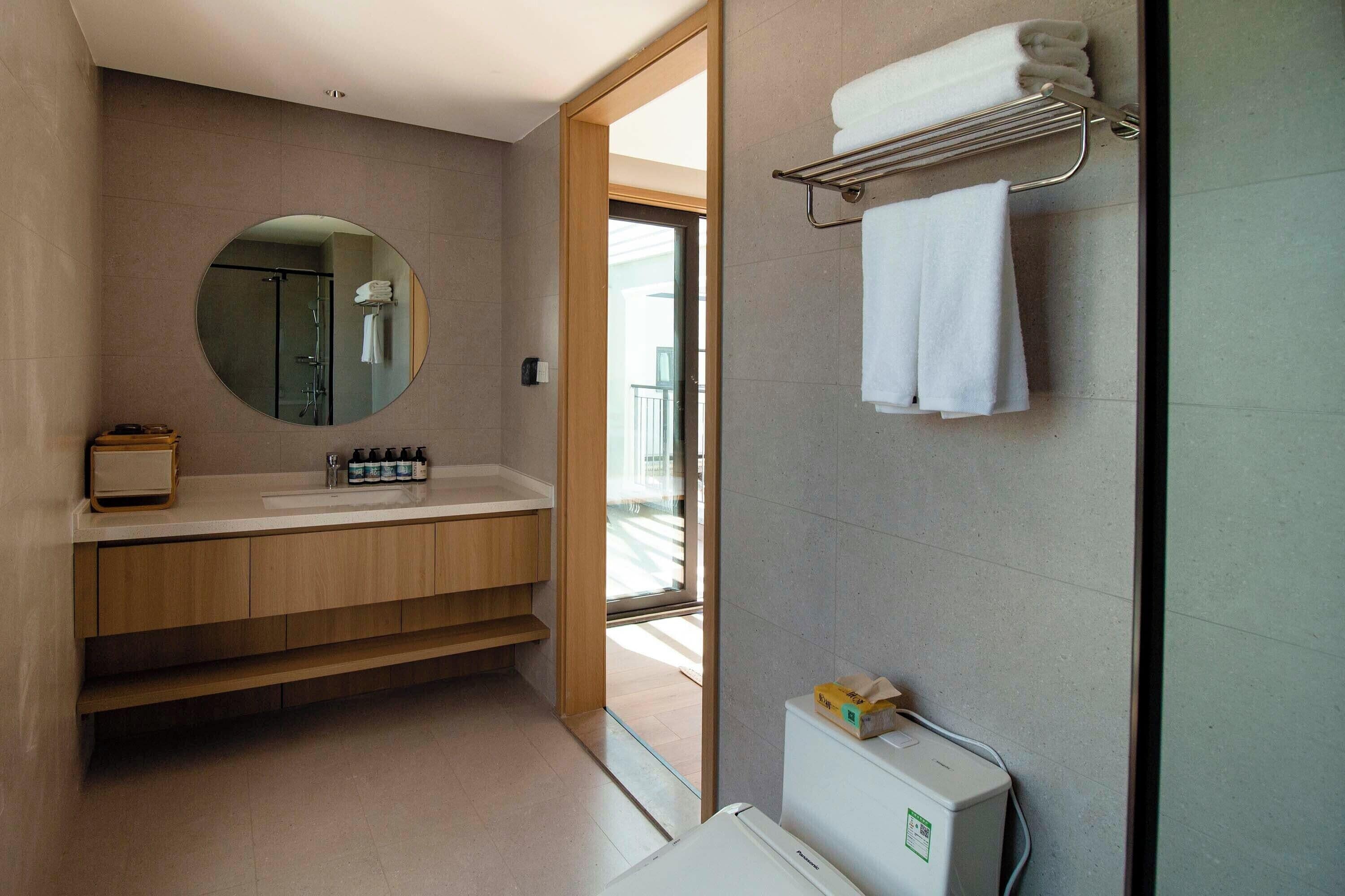 A modern bathroom with a wooden vanity, a round mirror, a towel rack, and a toilet with a visible water heater.