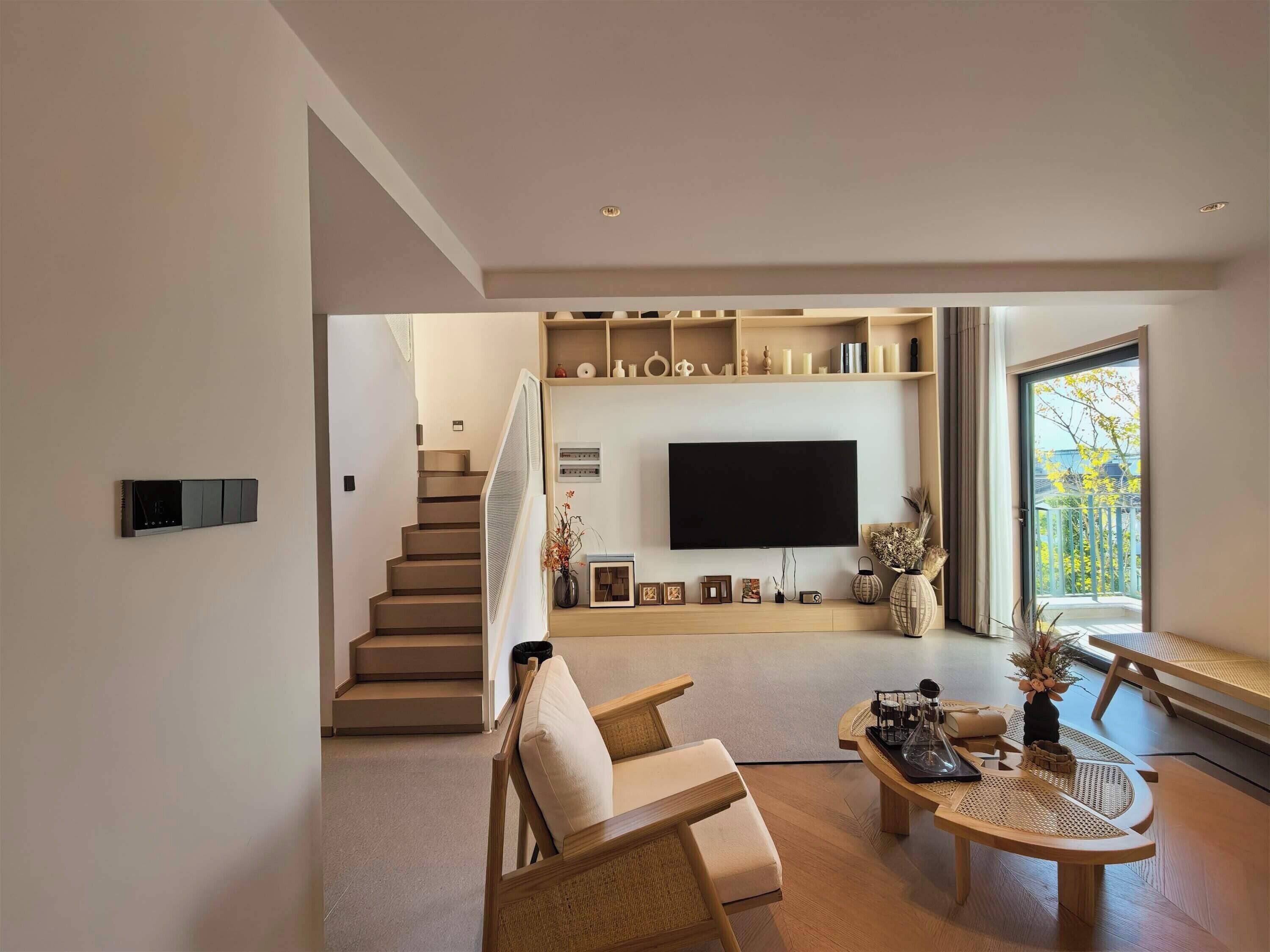 A modern living room with a staircase, a flat-screen TV, and a large window.