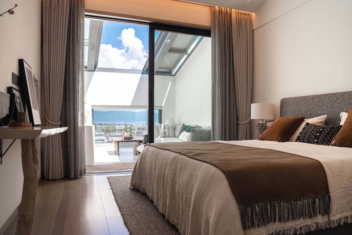 Quarto moderno com uma cama grande, mesa de cabeceira, luminária e vista da cidade através de uma porta de vidro.