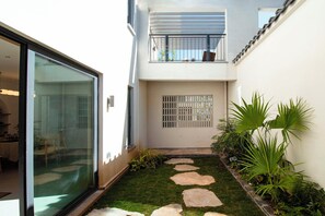 Un edificio moderno con puerta de cristal, un pequeño jardín con adoquines y un balcón con barandilla de metal.