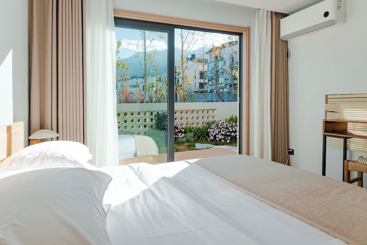 Quarto de hotel com uma cama grande, vista para um jardim e edifícios, e um aparelho de ar condicionado.