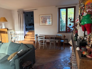 Ein Wohnzimmer mit einer Couch, einem Esstisch mit Stühlen, einer Treppe, einem Fenster mit Blick auf Bäume und einer Wand mit einem gerahmten Bild.
