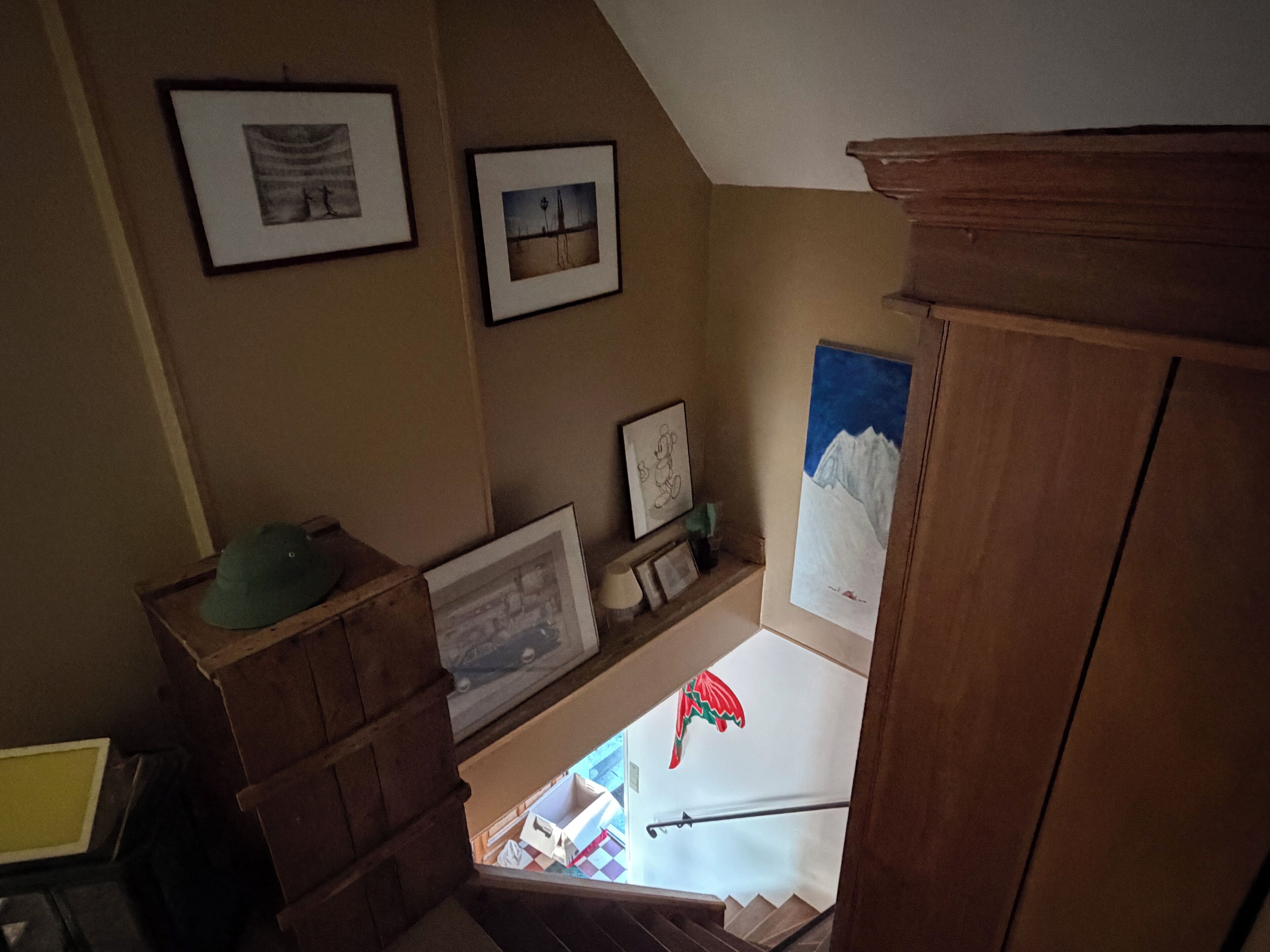 Un escalier avec une rampe en bois, un chapeau vert posé sur une boîte et des tableaux encadrés accrochés aux murs.