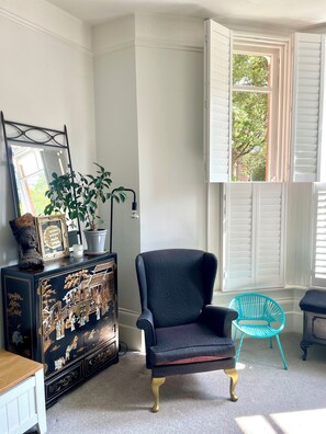 Una habitación con un sillón oscuro, una silla turquesa, un aparador decorativo y una ventana con contraventanas blancas.