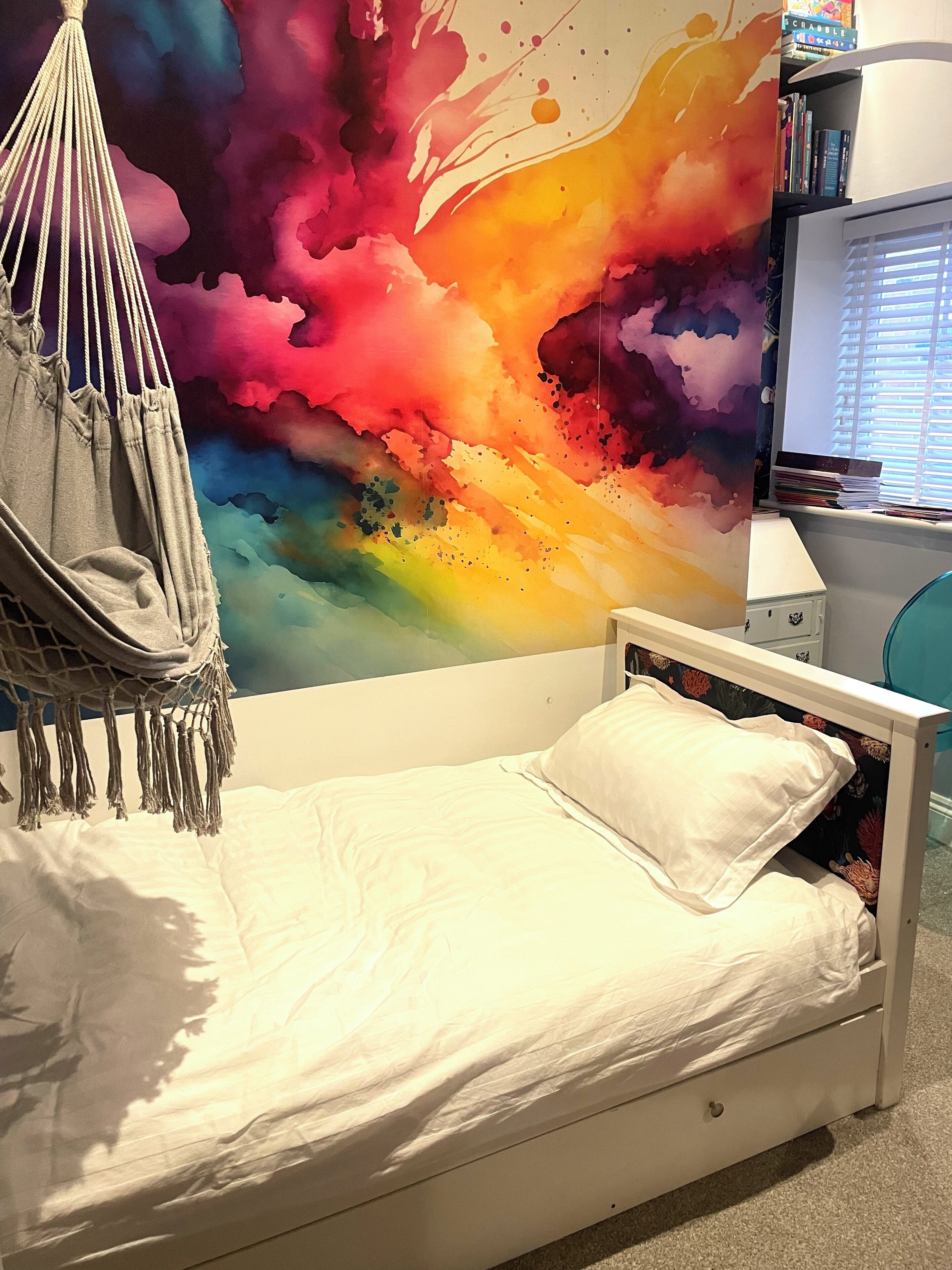 Un lit avec un matelas blanc et un seul oreiller blanc, un fauteuil suspendu gris et une peinture murale abstraite aux couleurs vives.