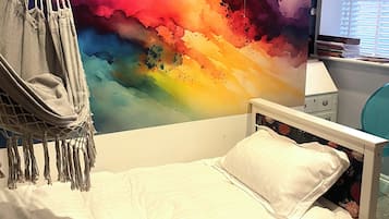 Un lit avec un matelas blanc et un seul oreiller blanc, un fauteuil suspendu gris et une peinture murale abstraite aux couleurs vives.