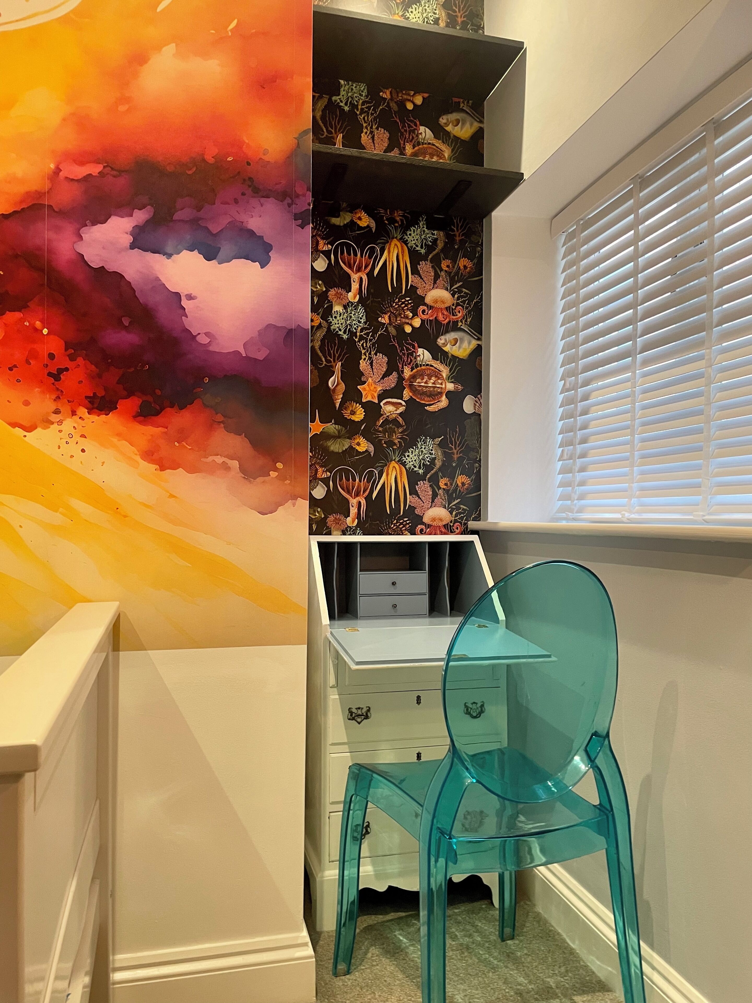 Ein Zimmer mit einem lebhaften Wandgemälde im abstrakten Stil, einem weißen Schreibtisch mit Schubladen, einem türkisblauen Sessel und einem Fenster mit Jalousien.