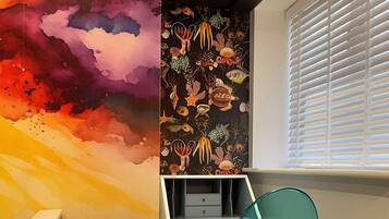Una habitación con un mural abstracto y llamativo en la pared, un escritorio blanco con cajones, una silla verde azulada y una ventana con persianas.