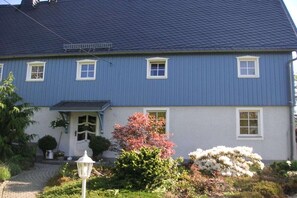 Ein zweigeschossiges Haus mit blauer Fassade und weißem Zierrat, einer Veranda und einem Garten mit verschiedenen Pflanzen und Blumen.