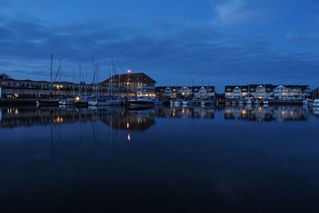 A marina with boats docked, a building with lights, and calm water reflecting the scene.