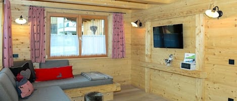 A cozy cabin interior with a sofa, two stools, a flat-screen TV, and a window with curtains.