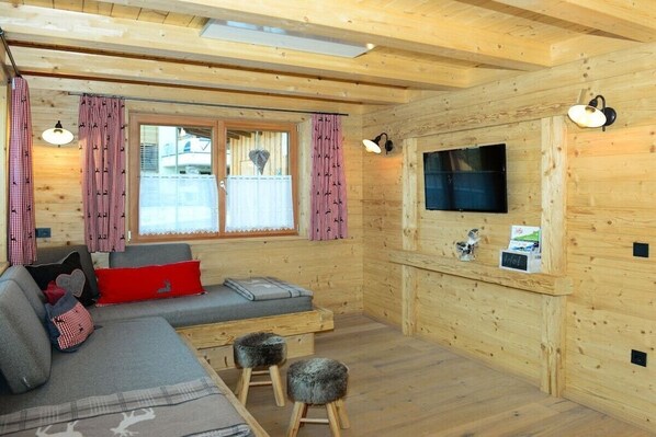 A cozy cabin interior with a sofa, two stools, a flat-screen TV, and a window with curtains.
