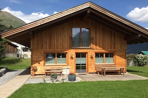 A wooden chalet with a balcony, large windows, and a deck with benches.
