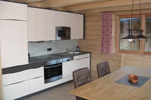 A compact kitchen with modern appliances, a dining area with a wooden table, and a window with patterned curtains.