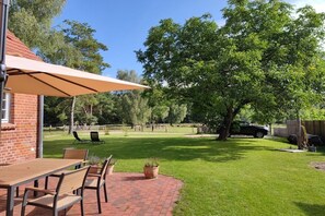 Un espace patio avec une table et des chaises en briques, un grand parasol, et une pelouse agrémentée d’arbres et d’une clôture.