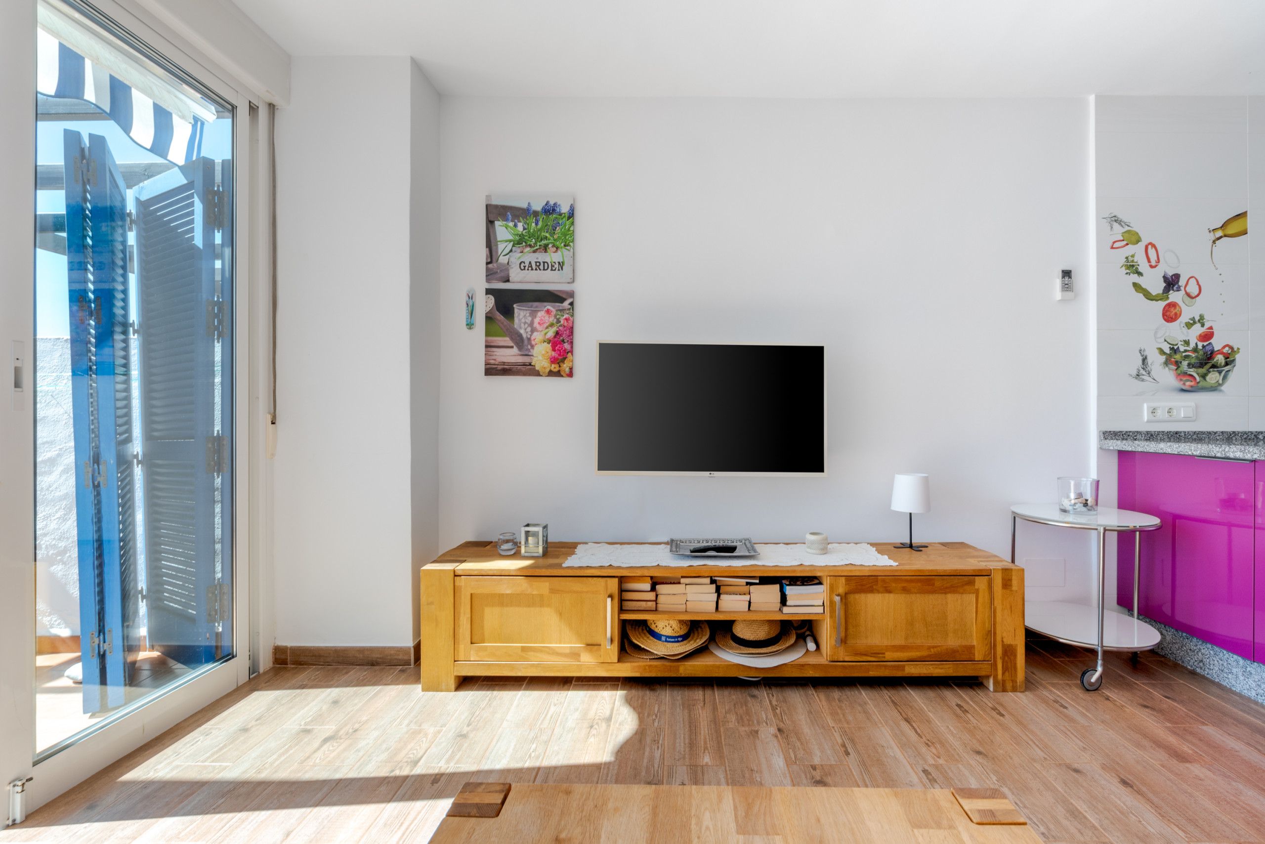 A modern living room with a wooden cabinet, a flat-screen TV mounted on the wall, and a large window providing a view of the cityscape.