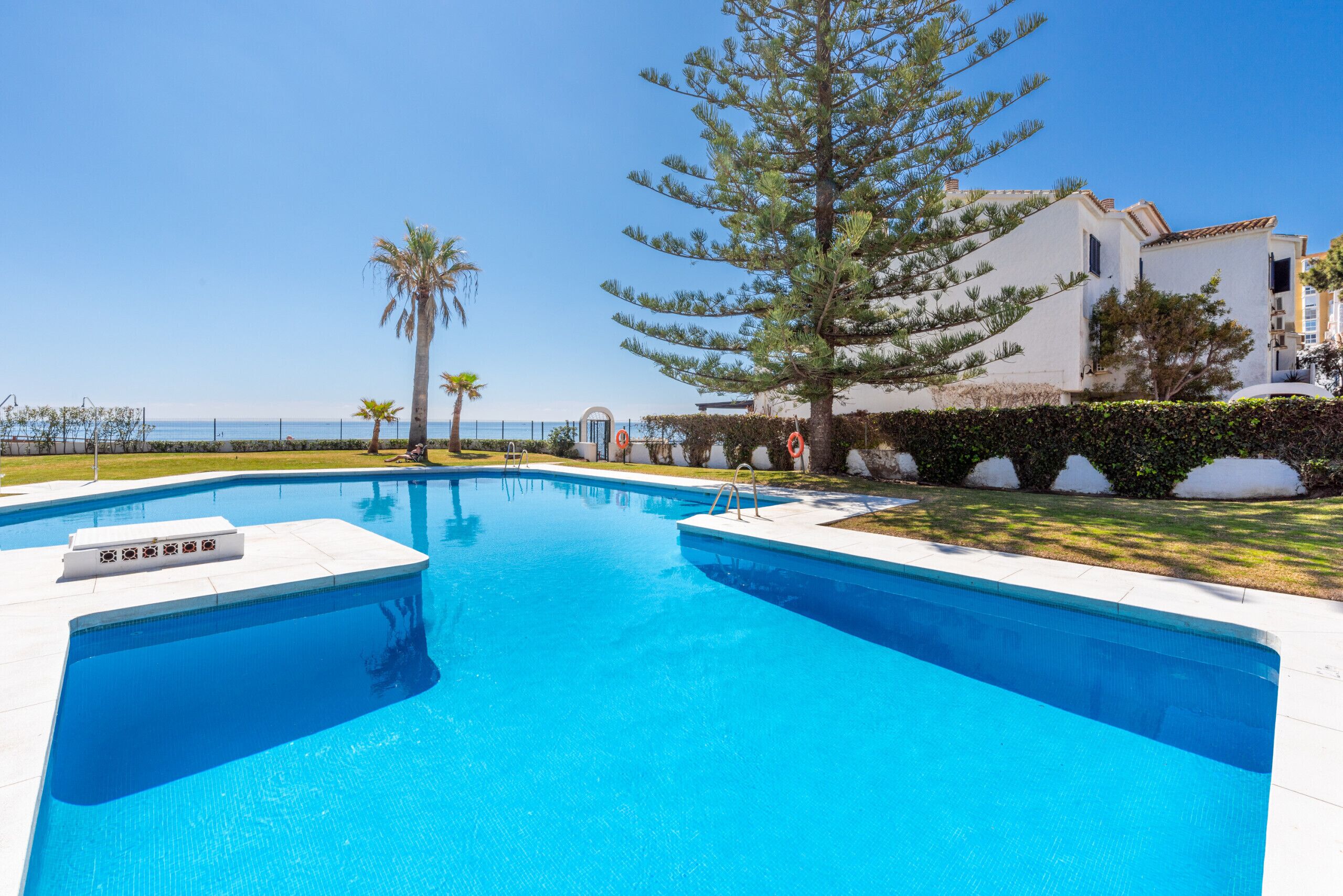 A swimming pool with a clear blue water, surrounded by a white pool deck, palm trees, and a building with a white facade.