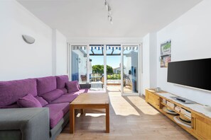 A living room with a purple sofa, a wooden coffee table, and a TV on a wooden stand. There is a sliding glass door leading to an outdoor patio with a table and chairs. - Calahonda Beach III – Beachfront Apartment with Sea Views in Mijas Costa (Mijas Costa)