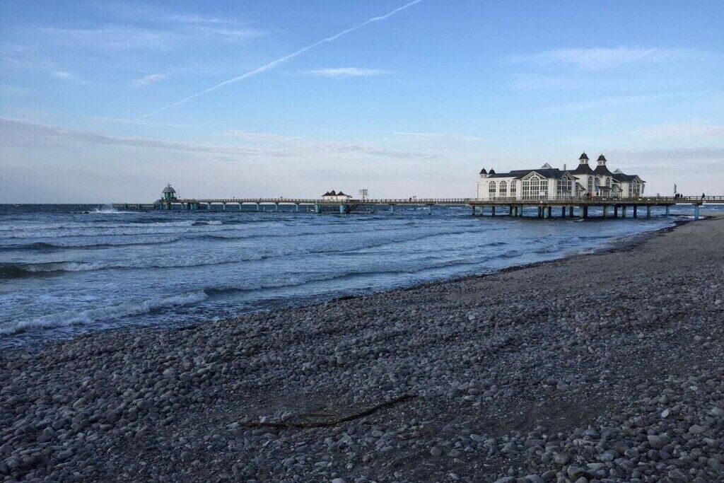 Ein Strand mit felsigem Ufer, einer in das Meer ragenden Promenade und einem großen Gebäude mit zahlreichen Fenstern und einem Turm.