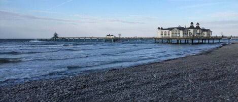 Une plage bordée d’un rivage rocheux, une jetée s’avançant dans la mer et un grand bâtiment doté de nombreuses fenêtres et d’une tour.