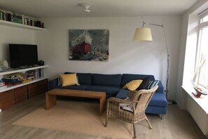 Uma sala de estar com sofá, mesa de centro, poltrona de vime, estante de livros, um quadro e abajur.