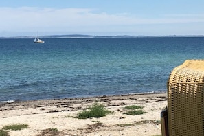 A beach with clear blue water, a sailboat in the distance, and a wicker chair on the sand. - Holiday home with 3 bedrooms (Döhnsdorf)