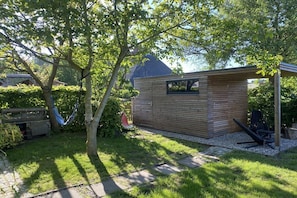 A wooden structure with a window, surrounded by a garden with a tree and a hammock. - Holiday home with 3 bedrooms (Döhnsdorf)