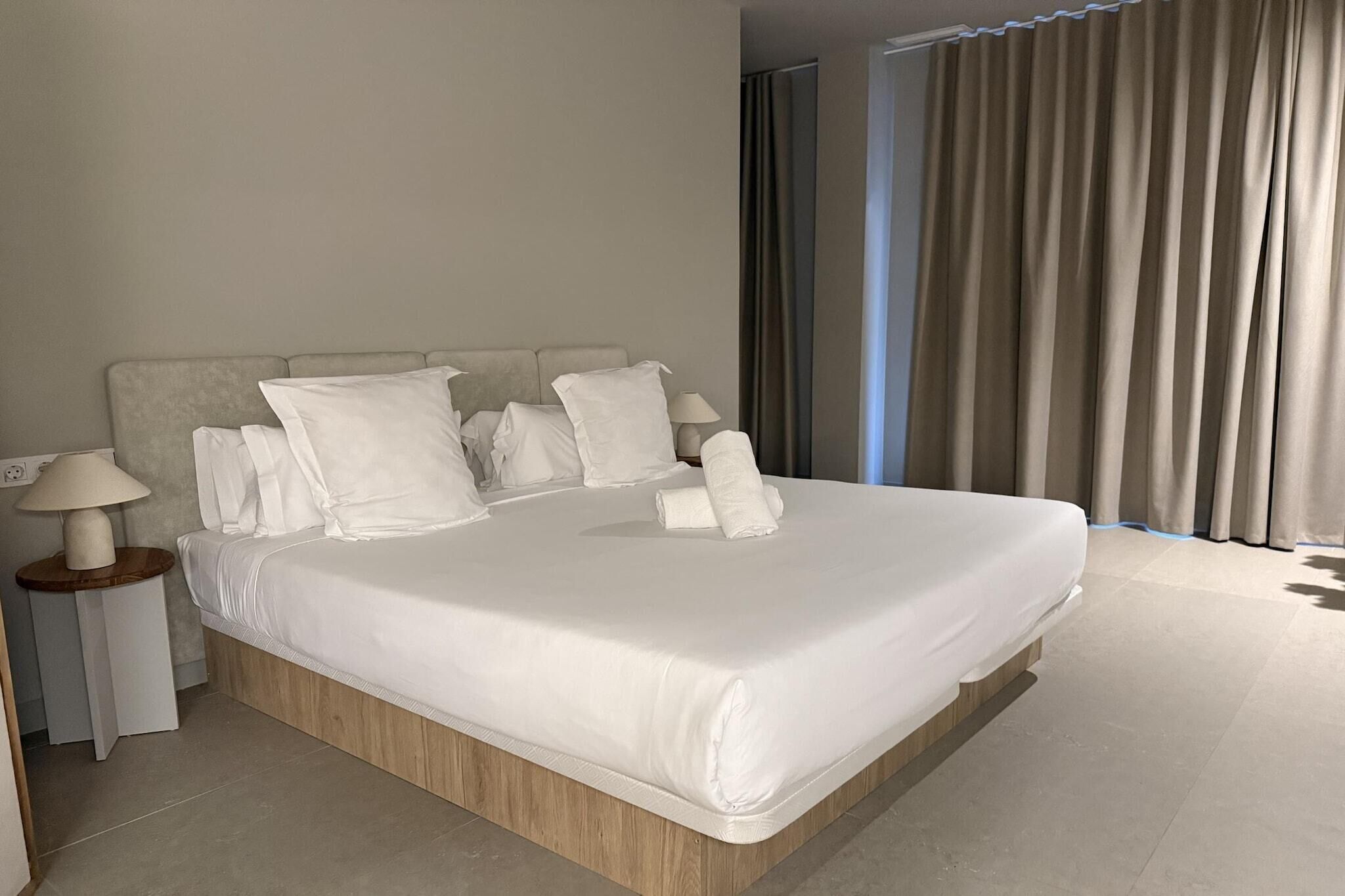 Une chambre d’hôtel moderne avec un grand lit, plusieurs oreillers, une table de chevet avec une lampe et une vue sur l’extérieur par une fenêtre munie de rideaux.