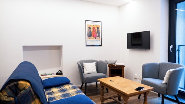 A modern living room with a blue sofa, a grey armchair, a wooden coffee table, and a small side table with a basket. A framed picture hangs on the wall, and a flat-screen TV is mounted above the armchair. - Brilliant apartment in Saxon for your getaway (Saxon)