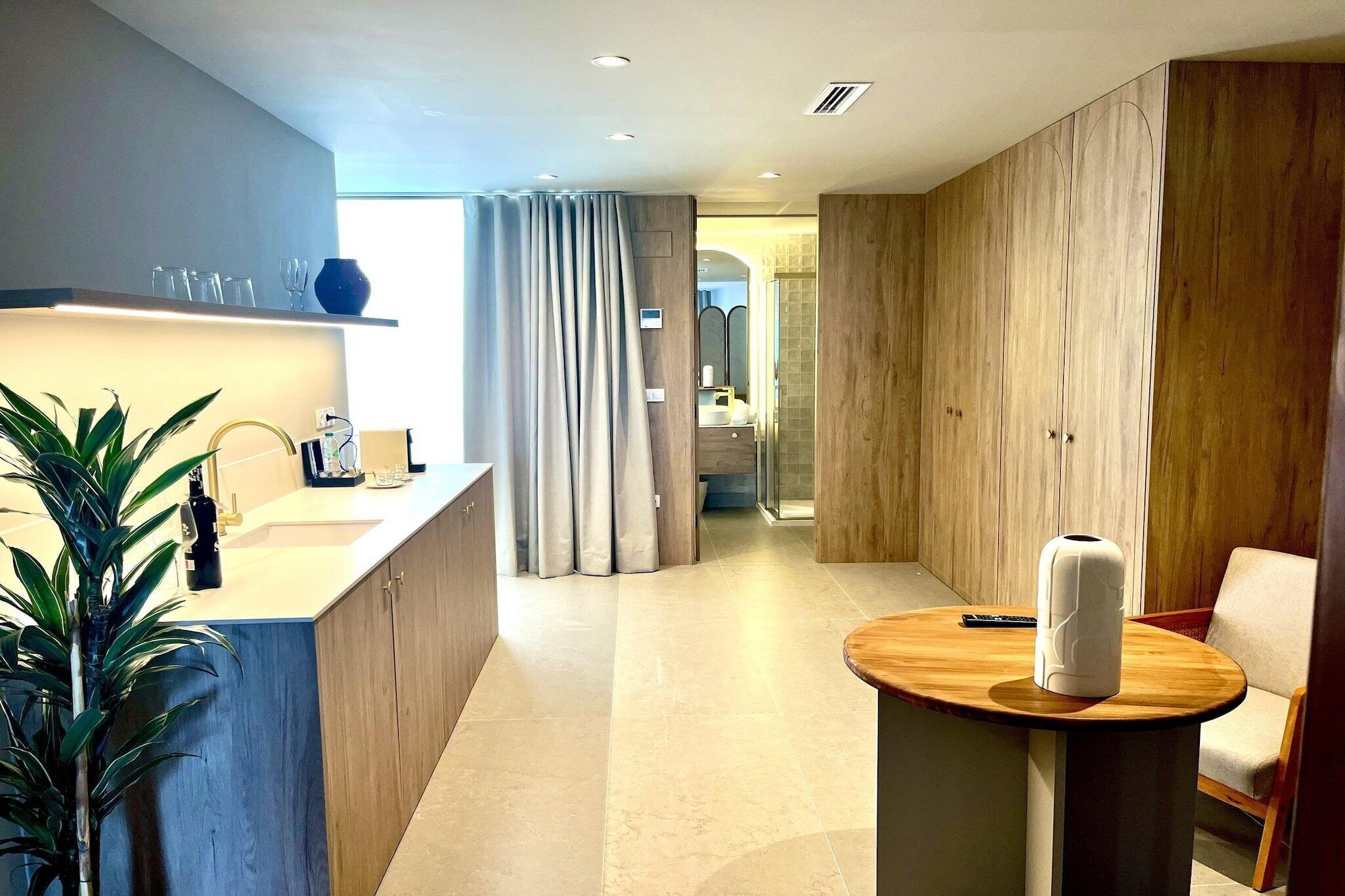 Et moderne hotelværelse med et træskab, et rundt træbord, en potteplante og et bruseområde.