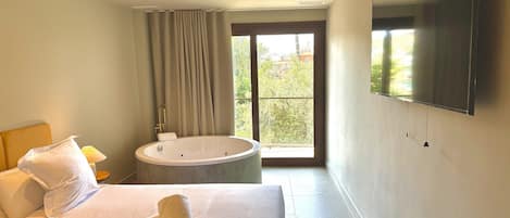 A hotel room with a large bed, a bathtub, a TV mounted on the wall, and a window with a view of greenery.