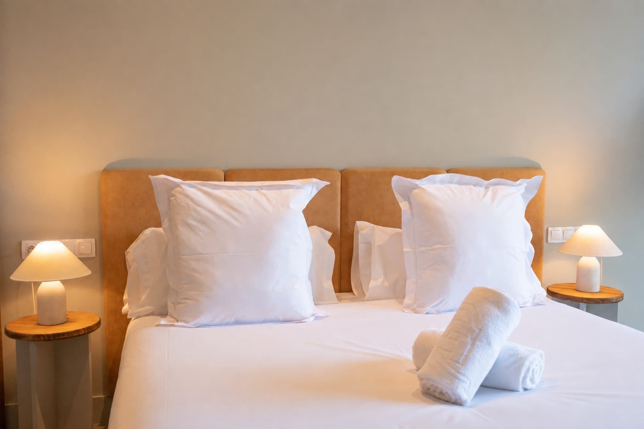 Een net opgemaakt bed met witte lakens en kussens, twee bedlampjes en een houten hoofdbord.