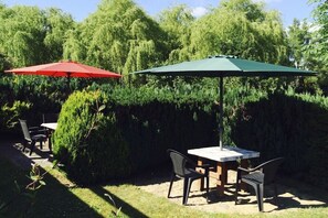 Eine Terrasse im Freien mit Tisch und Stühlen, zwei Sonnenschirmen und üppigem Grün im Hintergrund.