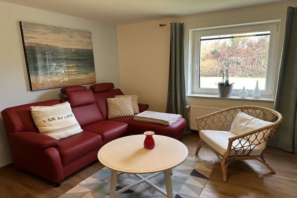 A living room with a red sofa, a wicker chair, a small round table, and a window with a view of trees.