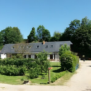 Een gebouw met twee verdiepingen, een donker dak en witte muren, omringd door groen en bomen.