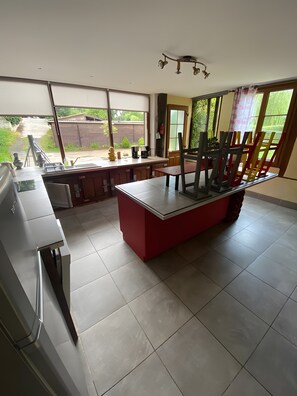 Een keuken met een centraal eiland, een eethoek met stoelen en grote ramen met uitzicht op de omgeving.