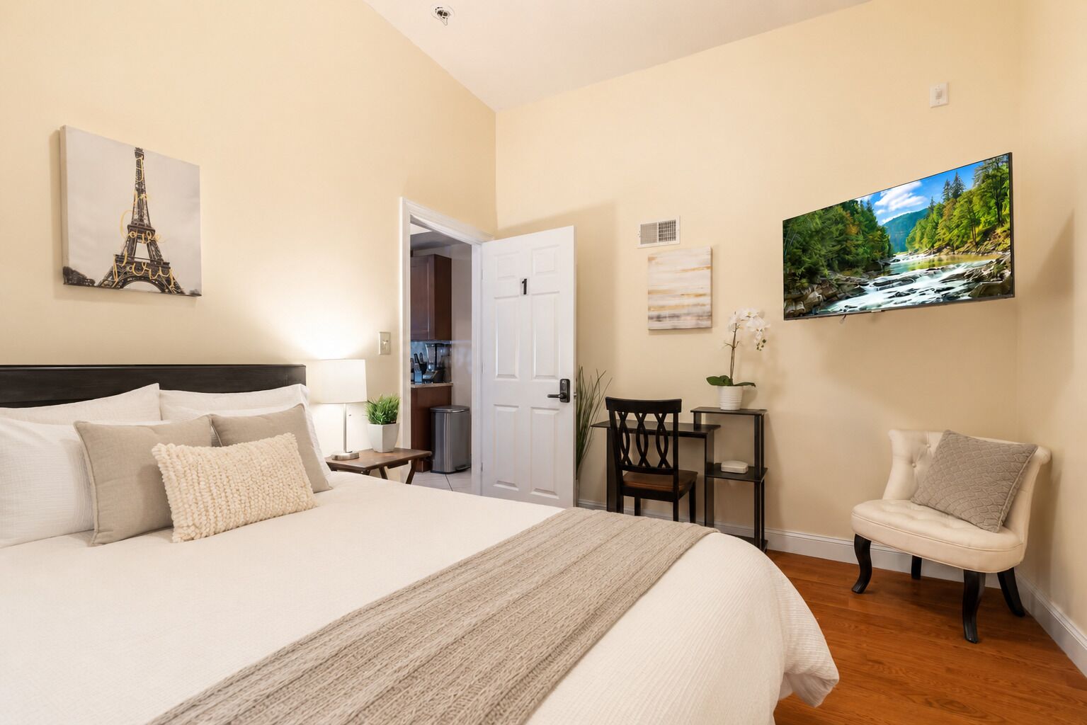 A bedroom with a large bed, a desk, a chair, a TV, and a door leading to a bathroom.